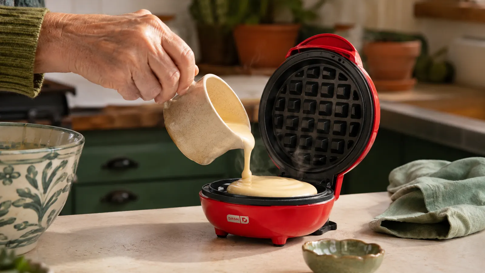 Hand pouring a small amount of waffle batter into the red DASH mini waffle maker on a countertop
