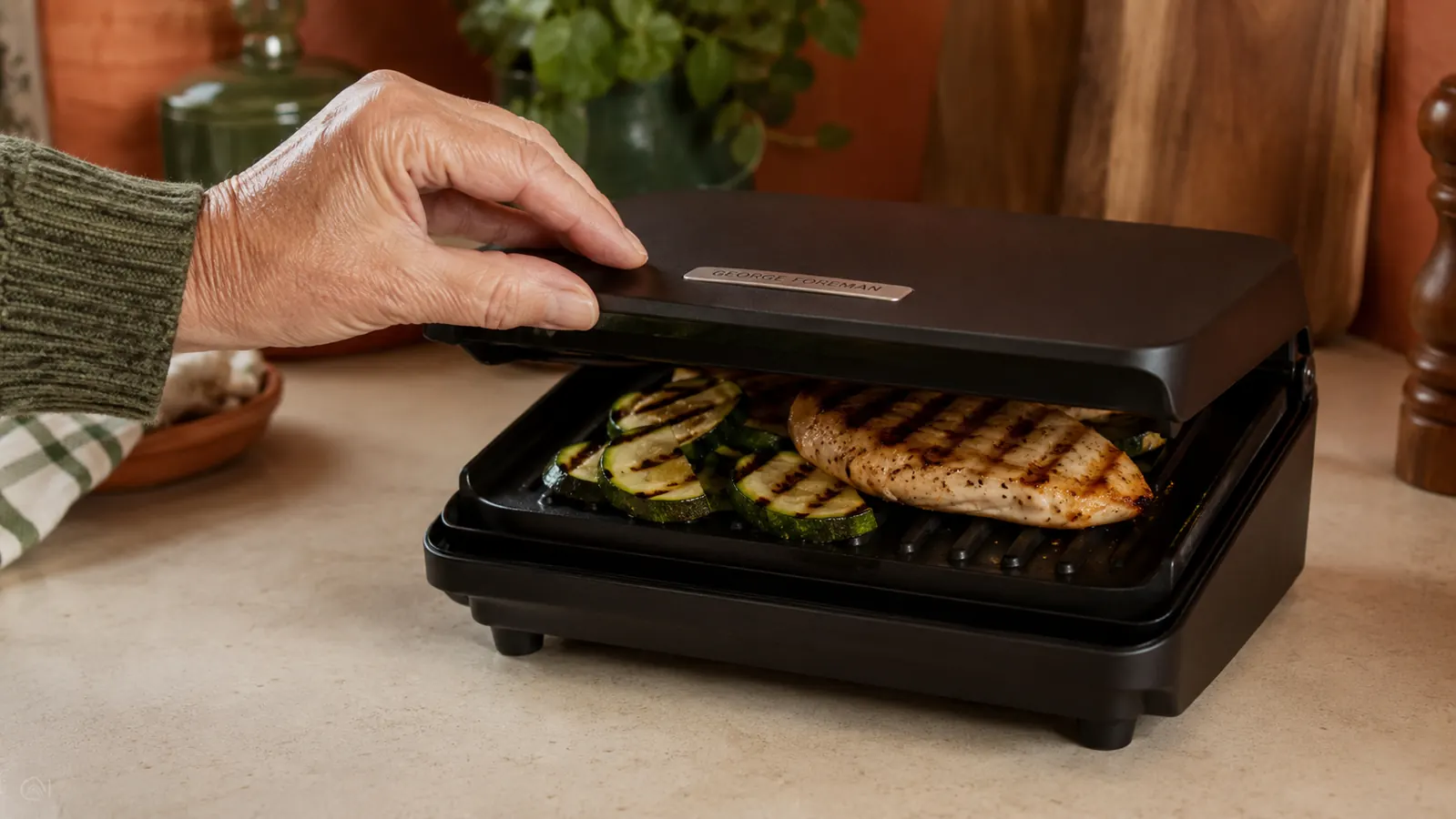 Hand lifting the top plate of the George Foreman mini grill to check a grilled chicken breast and vegetables