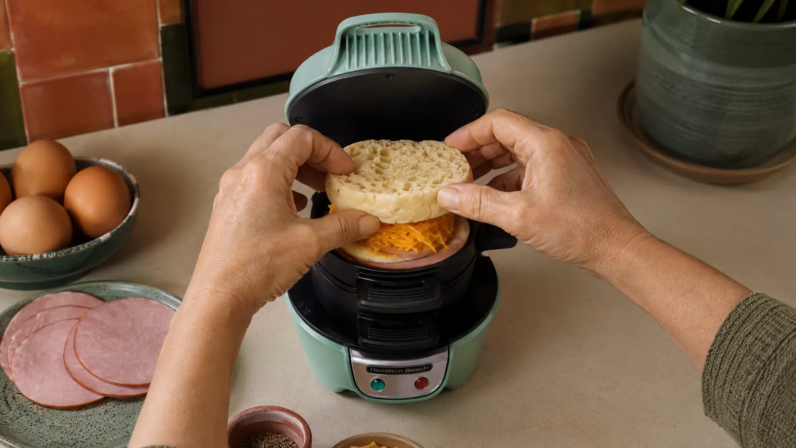 Hands loading an English muffin bottom, egg ring, and cheese slice into the layers of a Hamilton Beach breakfast sandwich maker