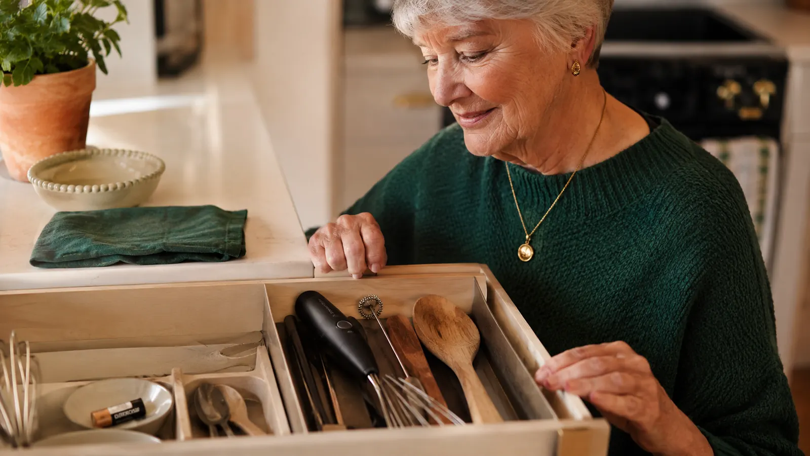 A small kitchen drawer opened to show the frother tucked among everyday utensils, next to a spare AA battery