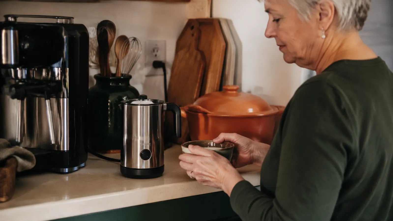 Standalone electric frother pitcher sitting on a crowded kitchen counter next to a coffee maker and toaster